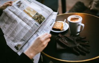 someone reading a newspaper with a piece of toast and a mug of coffee next to them