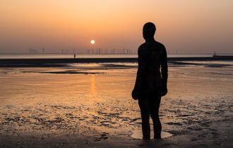 the sun sets over an offshore wind farm, in the foreground there is a beach with the tide out and an Anthony Gormley 'iron man' statue embedded in the sand One,Of,The,One,Hundred,Iron,Men,Statues,Which,Make
