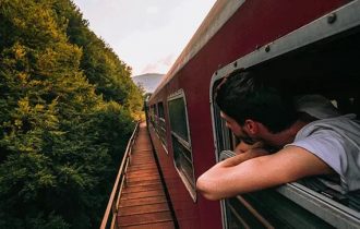 a man leaning out of a red train window as it runs alongside a hedge