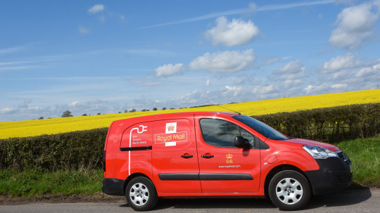 a red royal mail van driving past a yellow field on a sunny day