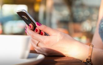 someone looking at a mobile phone - you can just see hands - next to a cup of something