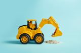 a yellow toy digger on a light blue background looks to be picking up a pile of grey dust