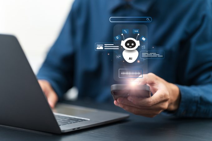 a man in a blue shirt is typing on a keyboard with one hand while holding a mobile in the other a white cartoon robot with Ai on it hovers above the mobile screen