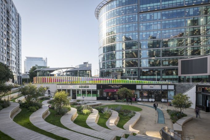 a view across the central area of paddngton central office complex, Sinuous steps picked out in wood with grass behind them step down to a curve of shops