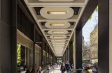 a view of Medici courtyard, a new square in Mayfair. The picture looks down a covered walkway with people sitting at metal tables. Above them, rows of oval lights stretch away into the distance. On the right there are trees and its a sunny day
