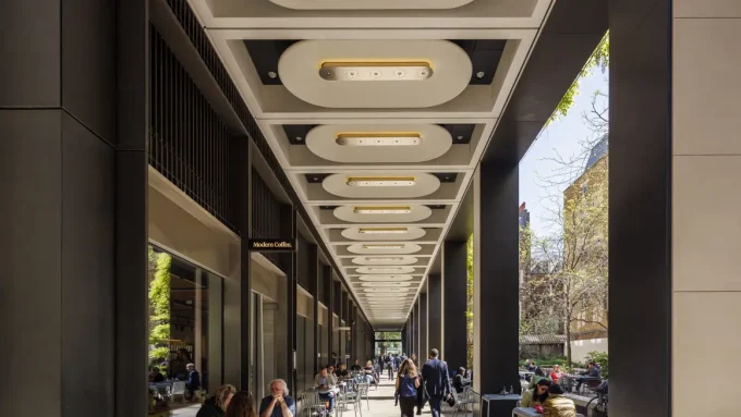 a view of Medici courtyard, a new square in Mayfair. The picture looks down a covered walkway with people sitting at metal tables. Above them, rows of oval lights stretch away into the distance. On the right there are trees and its a sunny day