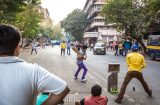 a group of people playing cricket in the street in mumbai, there's a taxi headed towards them