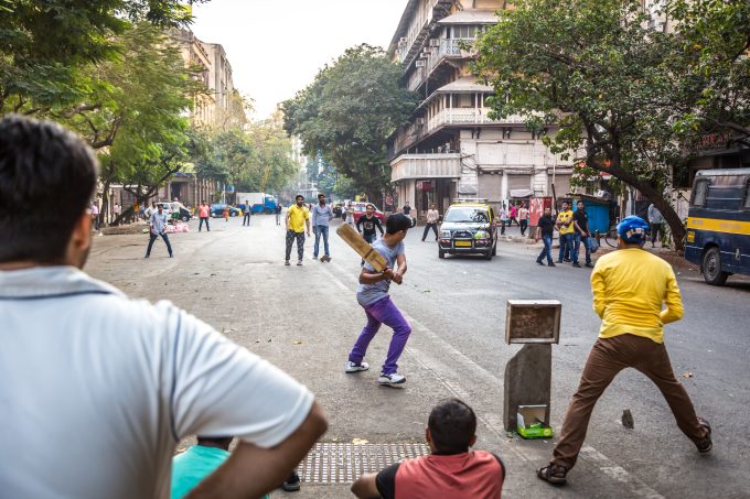 a group of people playing cricket in the street in mumbai, there's a taxi headed towards them